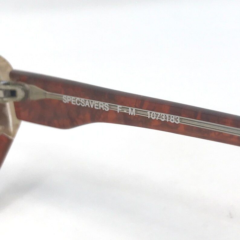 May include: Close-up of a brown and tan speckled eyeglass frame arm. The text "SPECSAVERS F-M 1073183" is printed on the arm in a silver color. The frame arm is made of a speckled brown material.