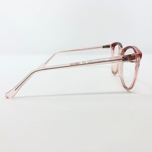May include: Pale pink eyeglasses with a cat-eye frame. The glasses have a translucent frame and temples, with visible text on the temple. The lenses are clear. The glasses are set against a white background.