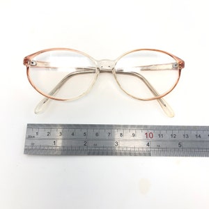 May include: Clear oval eyeglasses with a light brown gradient on the upper rims. The glasses are next to a ruler. The frame is a light, transparent colour. The ruler shows measurements in centimetres.
