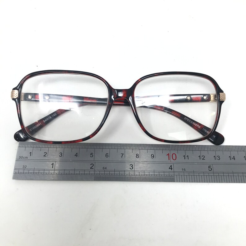 May include: Eyeglasses with a tortoiseshell pattern frame. The glasses have a rectangular shape and clear lenses. The frame has gold-colored accents on the temples. A ruler is placed below the glasses for measurement. The text "SPECSAVERS" is visible.
