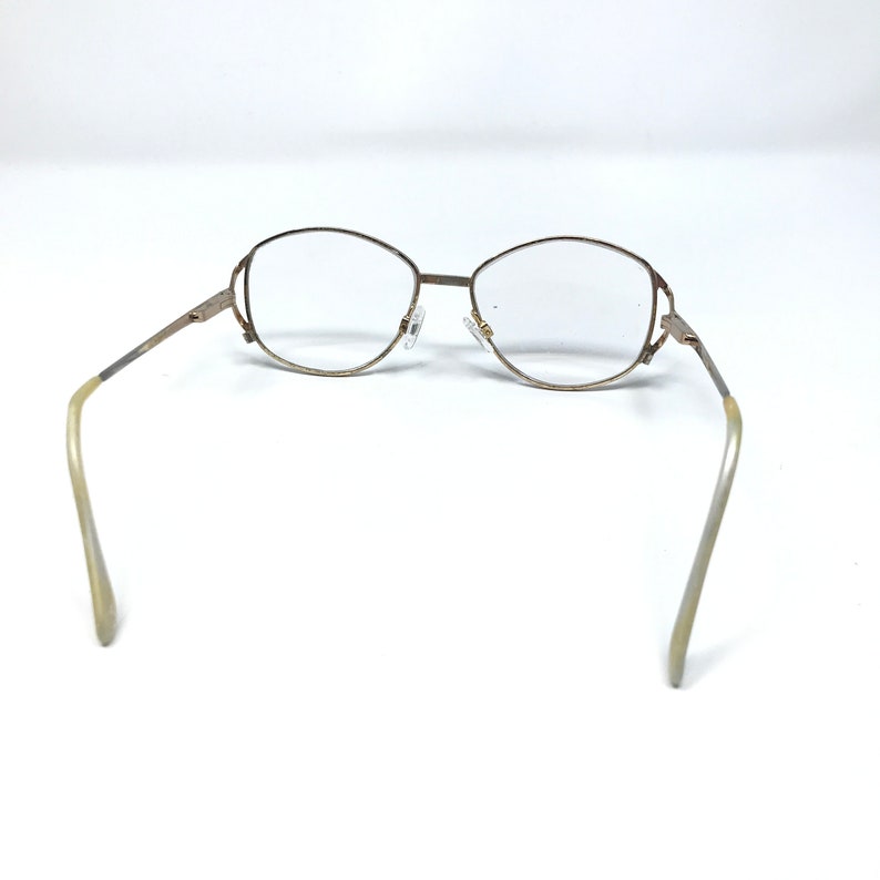 Op de afbeelding: Een bril met een goudkleurig metalen montuur en heldere glazen. De bril heeft een afgeronde ovale vorm en een dun montuur. De pootjes zijn lichtbeige van kleur. De bril staat tegen een witte achtergrond.