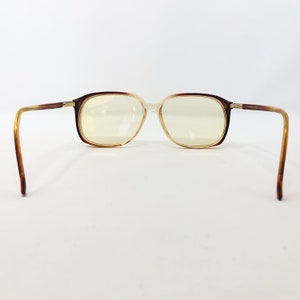 May include: Brown and tan eyeglasses with a rectangular frame. The lenses are a light yellow color. The arms of the glasses are a gradient of brown and tan. The glasses are set against a white background.