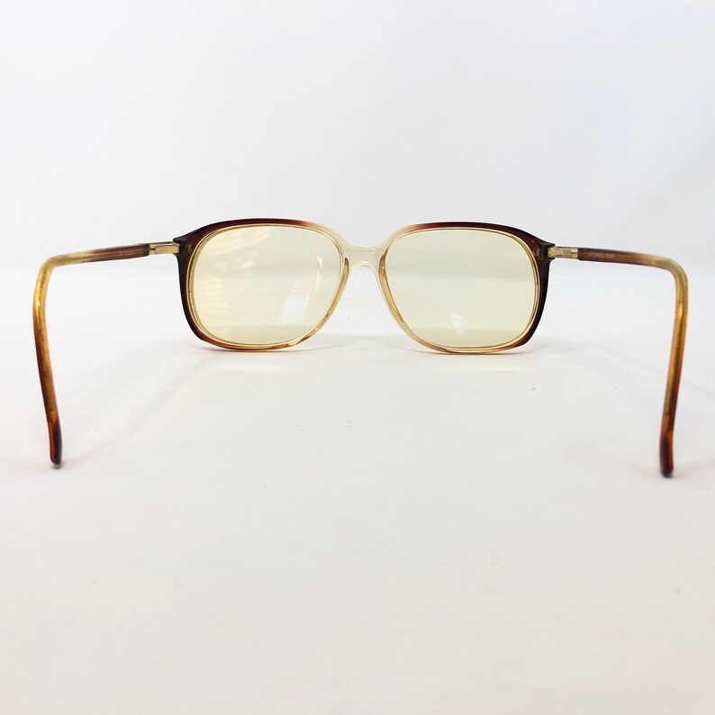 May include: Brown and tan eyeglasses with a rectangular frame. The lenses are a light yellow color. The arms of the glasses are a gradient of brown and tan. The glasses are set against a white background.