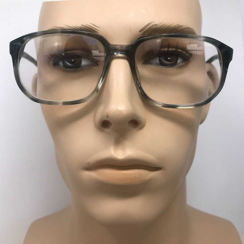 Puede incluir: Gafas de montura cuadrada con un patr&oacute;n veteado gris y transparente. Las gafas tienen una montura gruesa y lentes transparentes. Las gafas se exhiben en una cabeza de maniqu&iacute;.
