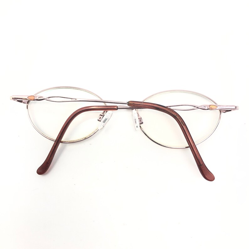 K&ouml;nnte beinhalten: Ovale Brille mit silberfarbenem Rahmen und braunen B&uuml;geln. Die Gl&auml;ser sind klar, und der Nasensteg ist silberfarben. Die B&uuml;gel biegen sich nach unten und au&szlig;en, vor wei&szlig;em Hintergrund.