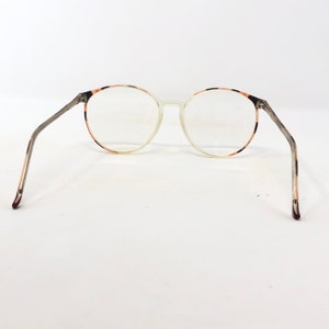 K&ouml;nnte beinhalten: Runde Brille mit klaren Gl&auml;sern und einem durchscheinenden Rahmen. Der Rahmen hat ein Schildpattmuster an den Au&szlig;enkanten und B&uuml;geln. Die B&uuml;gel sind eine Mischung aus Grau und Braun. Die Brille ist vor einem wei&szlig;en Hintergrund platziert.