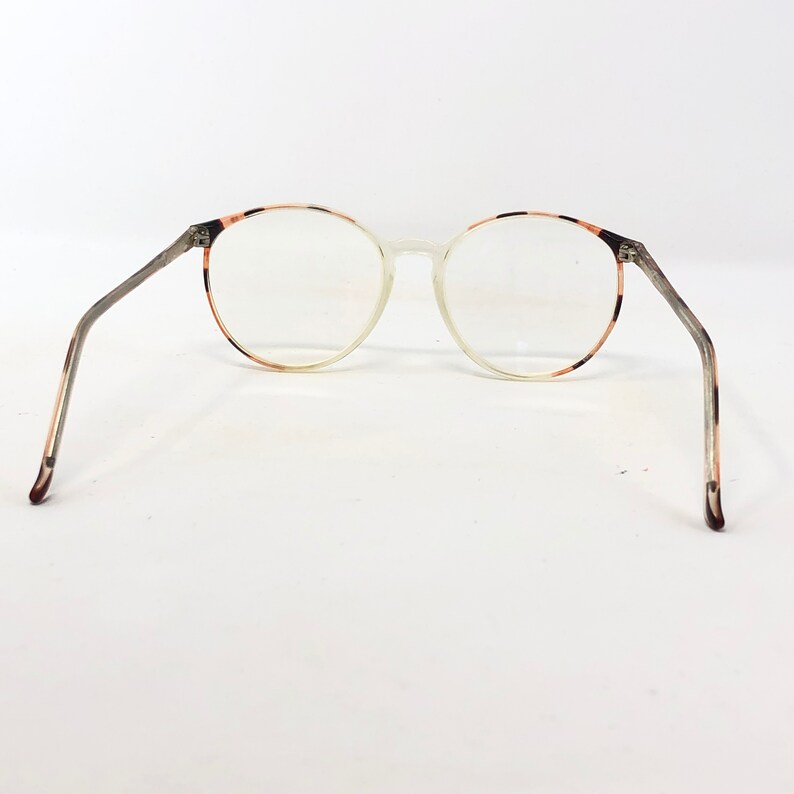 K&ouml;nnte beinhalten: Runde Brille mit klaren Gl&auml;sern und einem durchscheinenden Rahmen. Der Rahmen hat ein Schildpattmuster an den Au&szlig;enkanten und B&uuml;geln. Die B&uuml;gel sind eine Mischung aus Grau und Braun. Die Brille ist vor einem wei&szlig;en Hintergrund platziert.