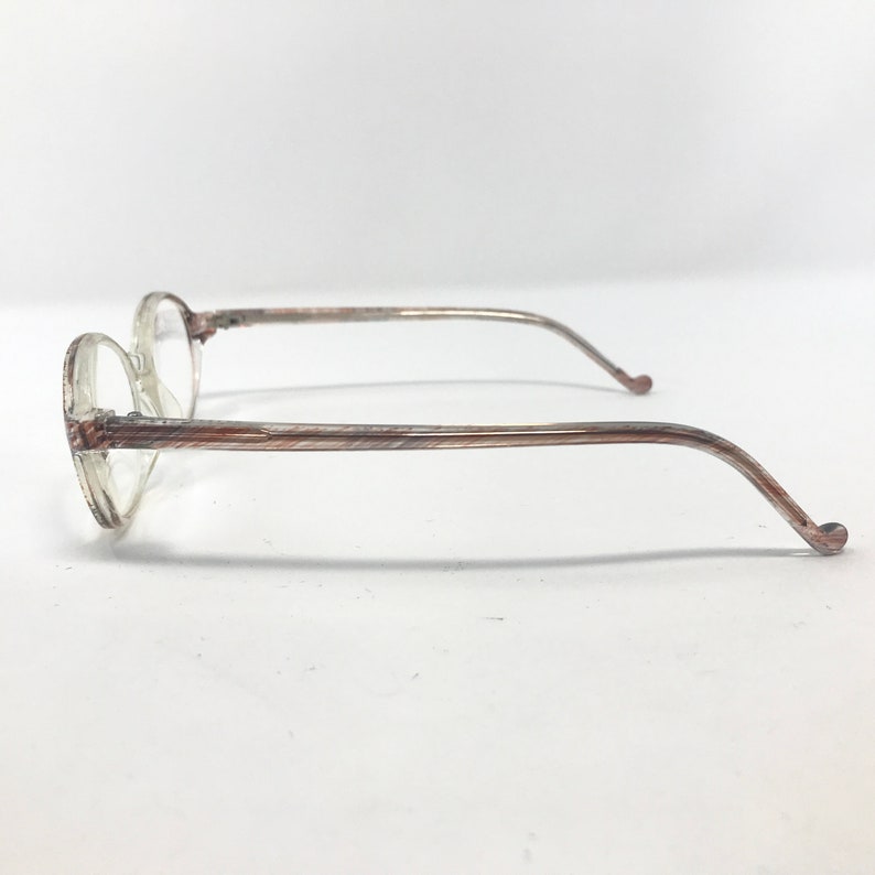 May include: Clear-framed spectacles with round lenses and brown and tan patterned temples. The side view shows the thin, curved arms extending from the frames. The glasses are set against a plain white background.