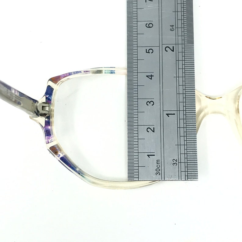 Puede incluir: Gafas transparentes con un patr&oacute;n decorativo en la parte superior de la montura, medidas con una regla. La montura tiene una bisagra plateada y una lente transparente. La regla muestra una medida de aproximadamente 10 cm.