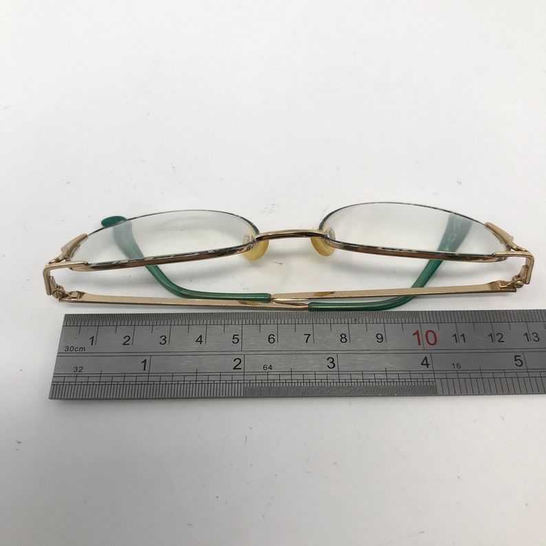 Pu&ograve; includere: Occhiali dorati con aste verdi e lenti trasparenti. Gli occhiali sono misurati con un righello, mostrando una lunghezza di 10 centimetri.