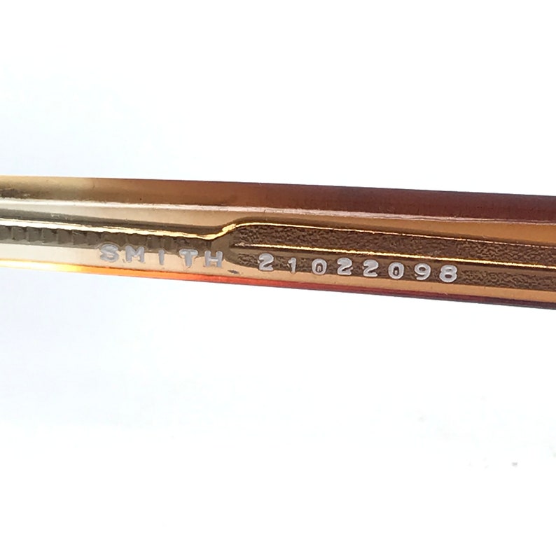 May include: Close-up of the brown and gold frame of a pair of glasses with the text "SMITH 21022098" printed on the inside of the frame.