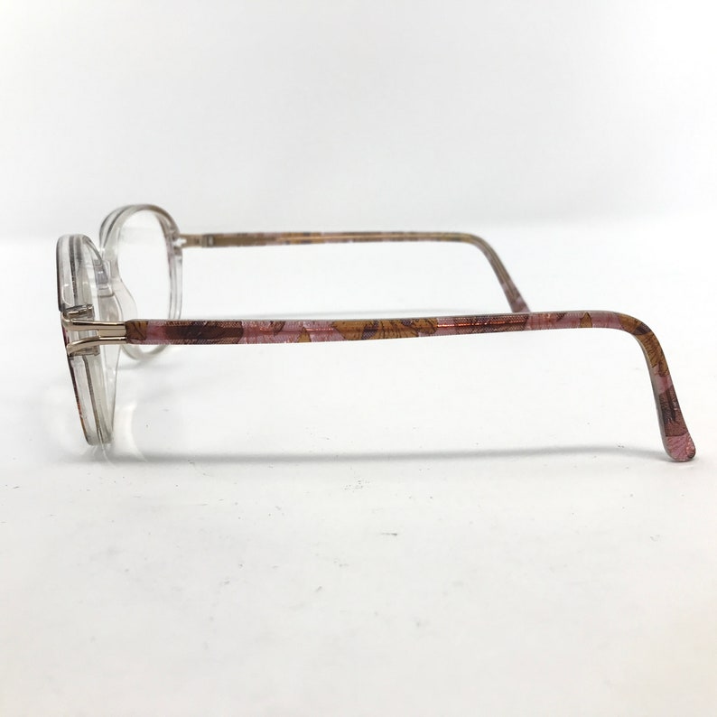 May include: Clear eyeglasses with a floral pattern on the temples. The frames are a transparent plastic, and the temples have a pink and brown floral design. The lenses are clear, and the glasses have a classic, understated style.