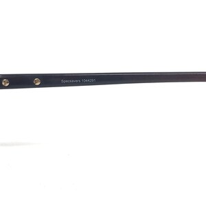 May include: A close-up of a black eyeglass frame with gold-coloured accents. The text "Specsavers 1044291" is printed on the frame. The frame is set against a white background.