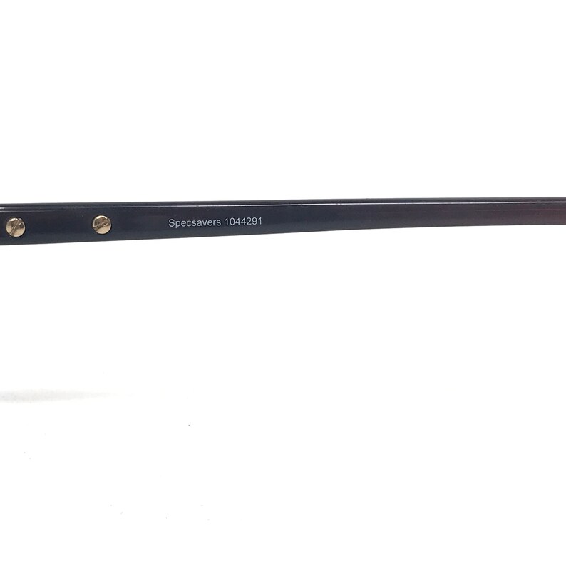 May include: A close-up of a black eyeglass frame with gold-coloured accents. The text "Specsavers 1044291" is printed on the frame. The frame is set against a white background.