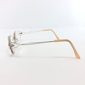 May include: Rimless eyeglasses with a silver metal frame and light beige temples. The lenses are oval-shaped and the frame has a delicate, minimalist design. The temples curve gently for a comfortable fit. The glasses are set against a white background.