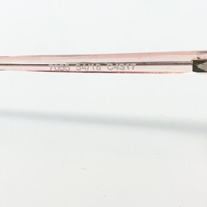 May include: A close-up of the arm of a pair of eyeglasses. The frame is a light pink color. The numbers "7035 54/16 C4317" are printed on the arm of the glasses.