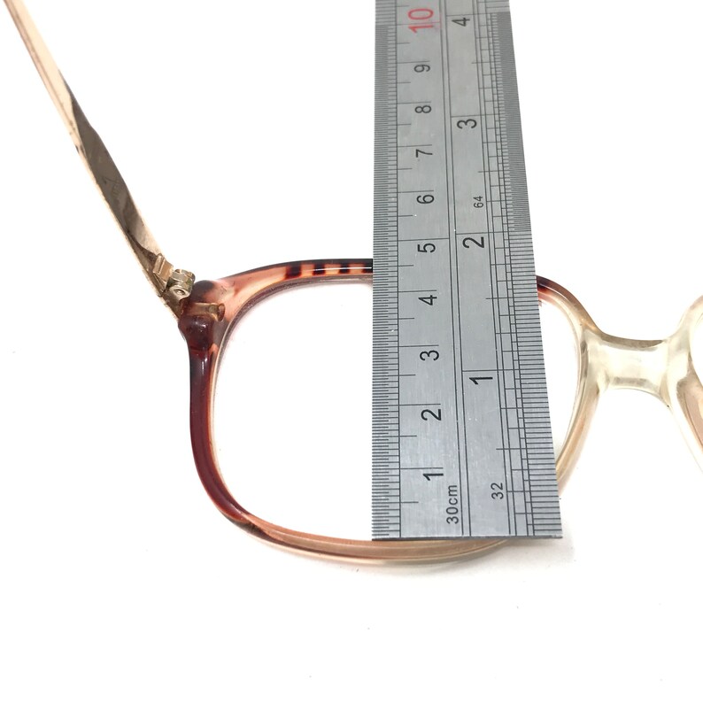 K&ouml;nnte beinhalten: Eine Brille mit braunen und klaren Gl&auml;sern und einem goldenen Metallrahmen. Die Brille wird mit einem Lineal gemessen, wobei die Breite des Rahmens 5,5 cm betr&auml;gt.