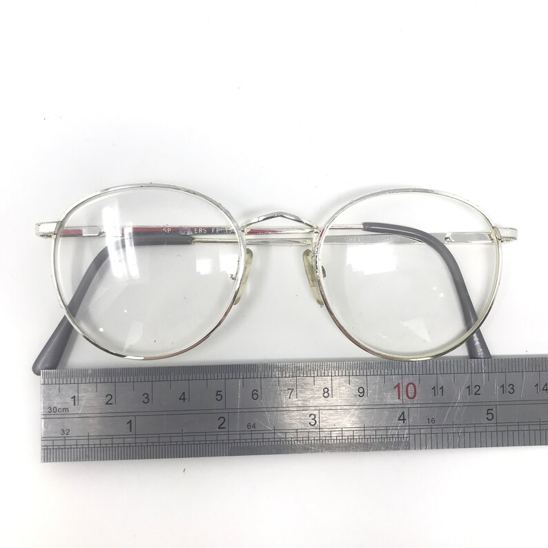 Peut inclure: Lunettes &agrave; monture argent&eacute;e avec verres ronds. Les lunettes ont des branches gris fonc&eacute; et un pont argent&eacute;. Une r&egrave;gle est plac&eacute;e en dessous, mesurant en centim&egrave;tres. Le texte "SPERS FP" est visible sur la monture.