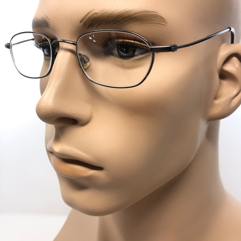 Puede incluir: Gafas con montura met&aacute;lica negra de forma rectangular redondeada. Las gafas est&aacute;n sobre una cabeza de maniqu&iacute;, mostrando el dise&ntilde;o y el ajuste de la montura. Las lentes son transparentes y las varillas son finas y elegantes.