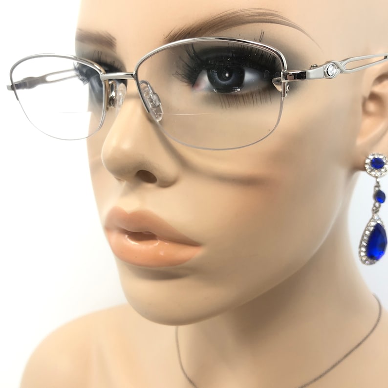 May include: Silver-toned eyeglasses with a half-rim design. The glasses have a delicate, curved frame and clear lenses. The image also shows a pair of blue teardrop earrings with a silver setting. The earrings are a vibrant blue color.