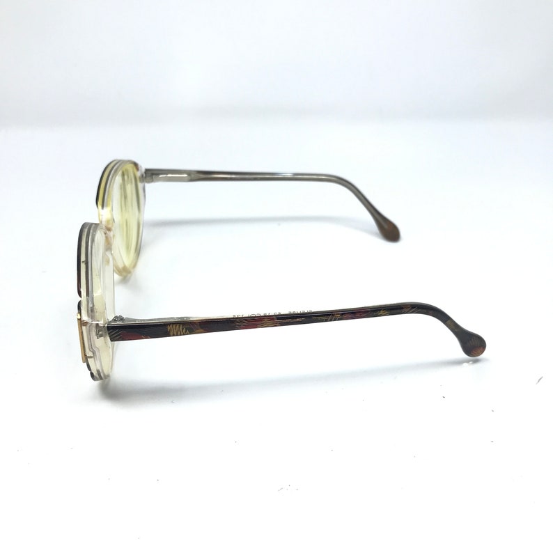 May include: A pair of vintage eyeglasses with round, clear frames and tinted lenses. The temples are a dark brown with a floral pattern, and the arms are a silver-gray color. The glasses are set against a white background.