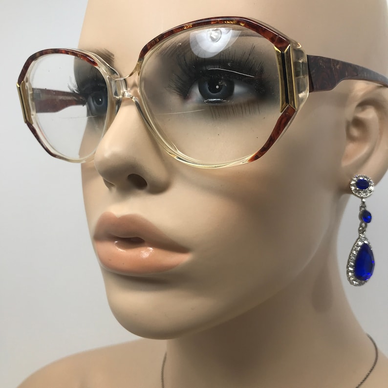 May include: Vintage eyeglasses with a clear frame and brown tortoise shell accents. The glasses have a geometric shape and are paired with dangling blue gemstone earrings. The glasses are on a mannequin.