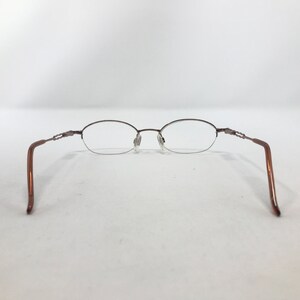 May include: Brown oval-shaped eyeglasses with a delicate metal frame. The glasses have a rimless design on the bottom half of the lenses and feature a decorative detail on the temples. The arms are a solid brown color.