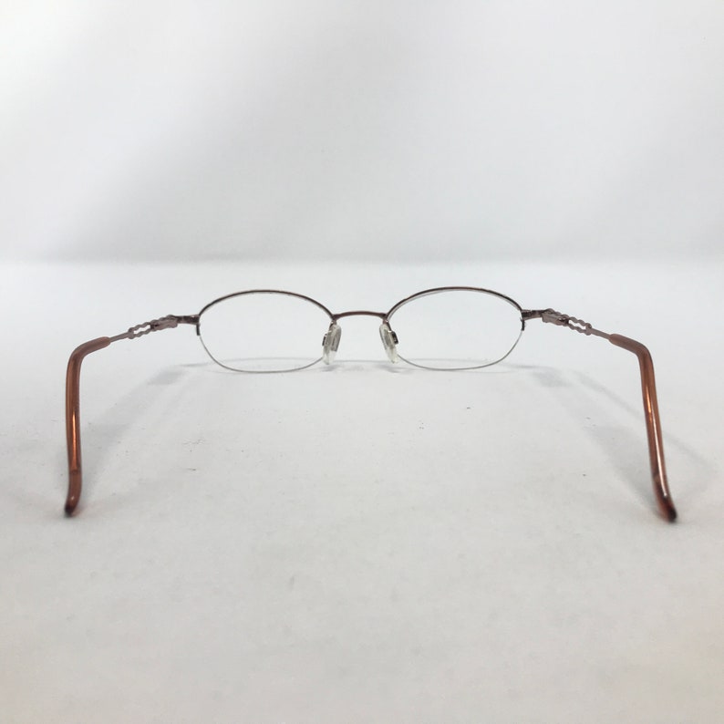 May include: Brown oval-shaped eyeglasses with a delicate metal frame. The glasses have a rimless design on the bottom half of the lenses and feature a decorative detail on the temples. The arms are a solid brown color.