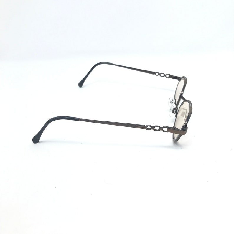 May include: A pair of oval-shaped eyeglasses with a dark brown metal frame. The temples and the frame around the lenses have a decorative chain-link design. The lenses are clear, and the nose pads are visible.