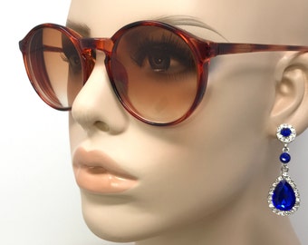 May include: Brown round sunglasses with gradient lenses and a pair of dangling blue and silver earrings. The earrings feature a small round blue stone at the top, a small blue stone in the middle, and a large teardrop-shaped blue stone at the bottom.