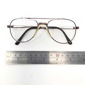 May include: Aviator-style eyeglasses with a dark brown metal frame and clear lenses. The glasses have a double bridge and a nose pad. The frame is approximately 5.5 inches wide. The text on the frame reads "140 Horack 21051023".