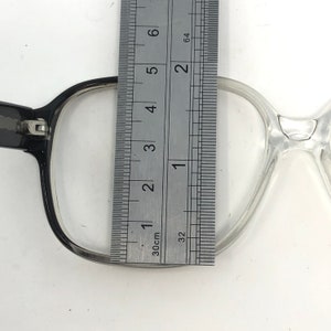 K&ouml;nnte beinhalten: Nahaufnahme einer Brille mit einem Lineal, das die Linsenbreite misst. Die Brille hat einen schwarzen und transparenten Rahmen. Das Lineal zeigt eine Linsenbreite von ca. 5 cm an.