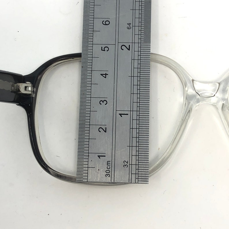 K&ouml;nnte beinhalten: Nahaufnahme einer Brille mit einem Lineal, das die Linsenbreite misst. Die Brille hat einen schwarzen und transparenten Rahmen. Das Lineal zeigt eine Linsenbreite von ca. 5 cm an.