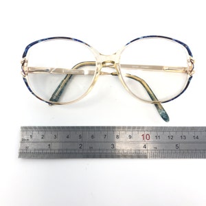 May include: Vintage eyeglasses with a clear frame and gold-tone metal accents. The upper rims feature a blue and white patterned design. The glasses are placed on a white surface next to a ruler.