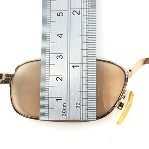 May include: Gold-framed sunglasses with brown lenses. The glasses are measured with a ruler, showing a width of approximately 2 inches. The lenses are a light brown color, and the frame is a shiny gold.