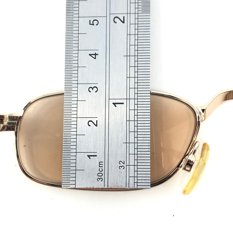 May include: Gold-framed sunglasses with brown lenses. The glasses are measured with a ruler, showing a width of approximately 2 inches. The lenses are a light brown color, and the frame is a shiny gold.