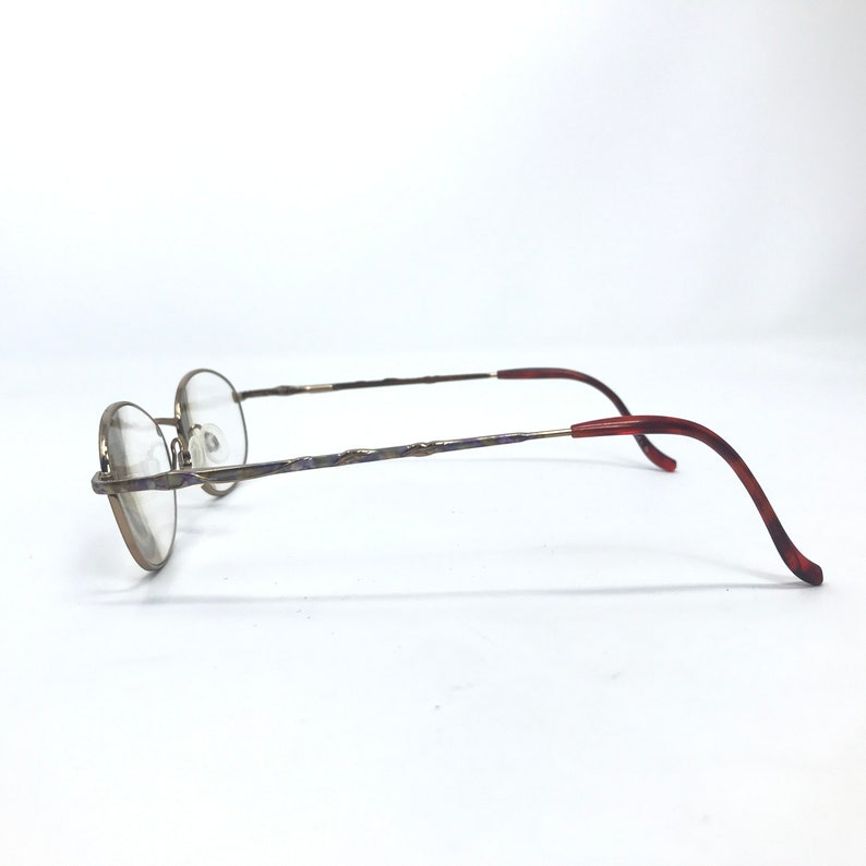 May include: A pair of gold-toned spectacles with a purple and gold patterned frame and red plastic temple tips.