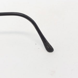 Puede incluir: Primer plano de una patilla de gafas negra. El brazo se curva suavemente y se estrecha hasta una punta redondeada. La superficie tiene un aspecto ligeramente texturizado y desgastado, lo que sugiere uso. El fondo es blanco liso.