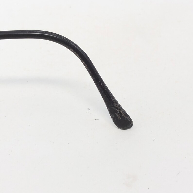 Puede incluir: Primer plano de una patilla de gafas negra. El brazo se curva suavemente y se estrecha hasta una punta redondeada. La superficie tiene un aspecto ligeramente texturizado y desgastado, lo que sugiere uso. El fondo es blanco liso.