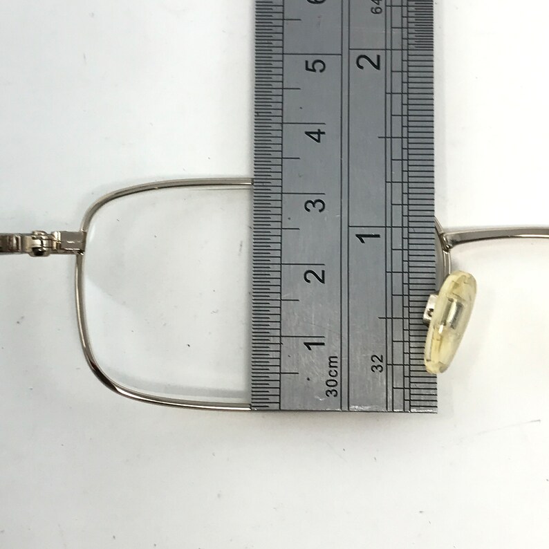 May include: Silver-framed eyeglasses with clear lenses are measured with a metal ruler. The ruler shows measurements in inches and centimeters. The glasses have a nose pad and a hinge.