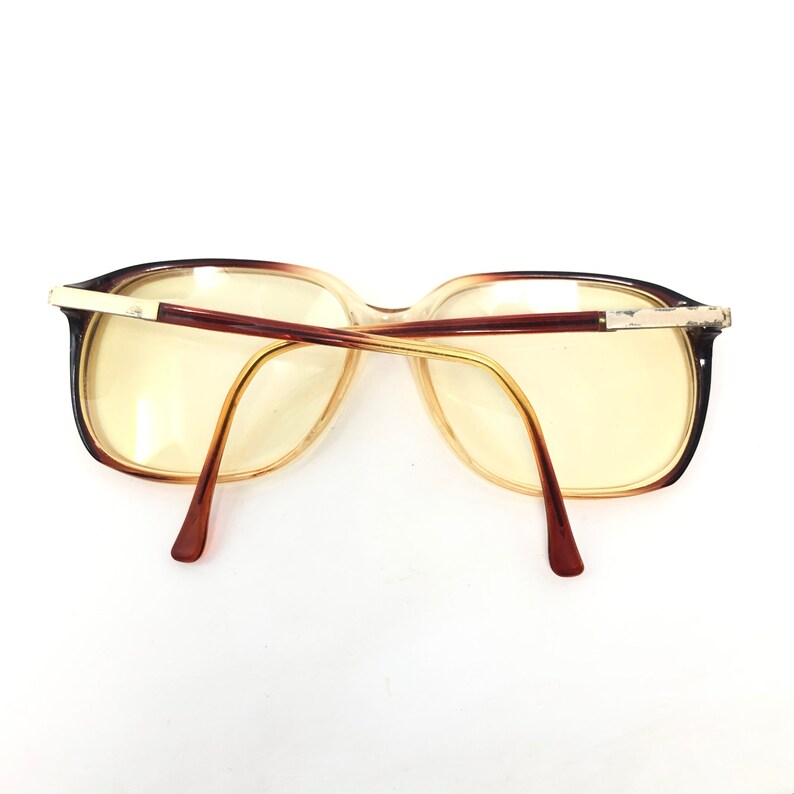 May include: Vintage eyeglasses with a brown and gold frame. The lenses are a light yellow color. The arms are a dark brown color and the frame has a rectangular shape.