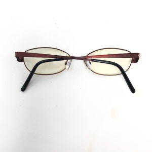 May include: Oval-shaped eyeglasses with a reddish-brown metal frame and black temples. The lenses are clear, and the nose pads are transparent. The glasses are designed for vision correction and are suitable for everyday use.