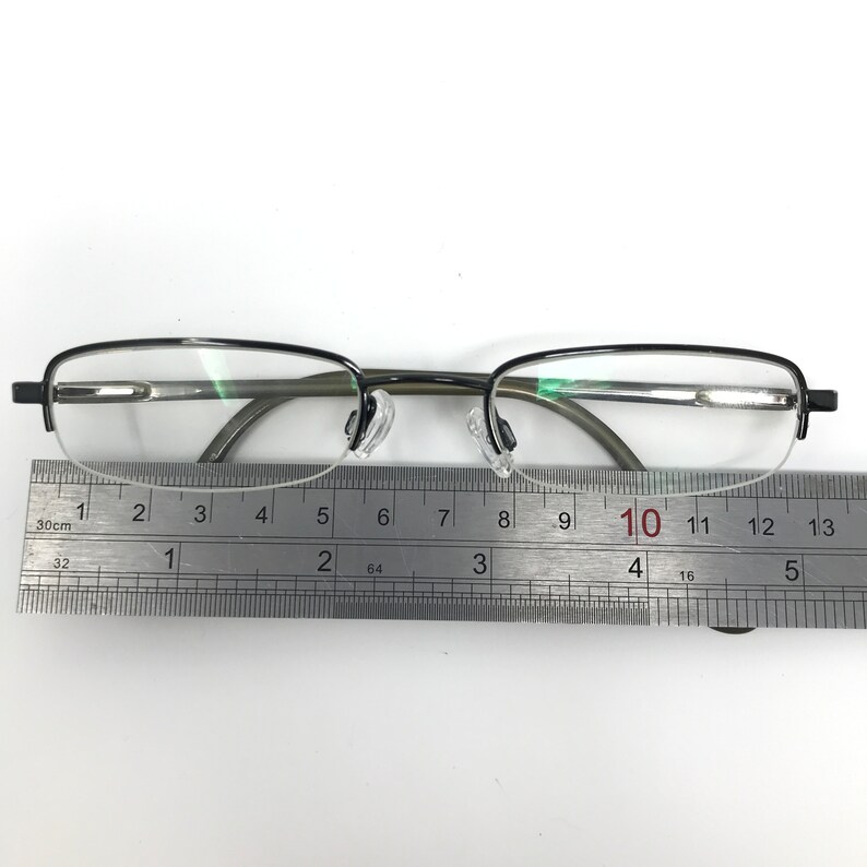K&ouml;nnte beinhalten: Halbrandbrille mit dunklem Metallrahmen und klaren Gl&auml;sern. Ein Metalllineal liegt darunter und zeigt die Ma&szlig;e in Zentimetern an. Die Brille hat ein klassisches, schlichtes Design.