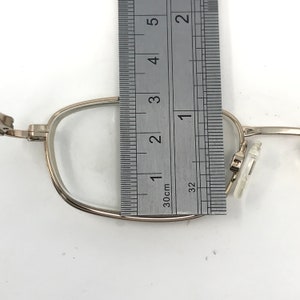 May include: Silver-framed eyeglasses with clear lenses are measured with a ruler. The frame is approximately 1 inch wide. The glasses are set against a white background, showing the details of the frame and lenses.