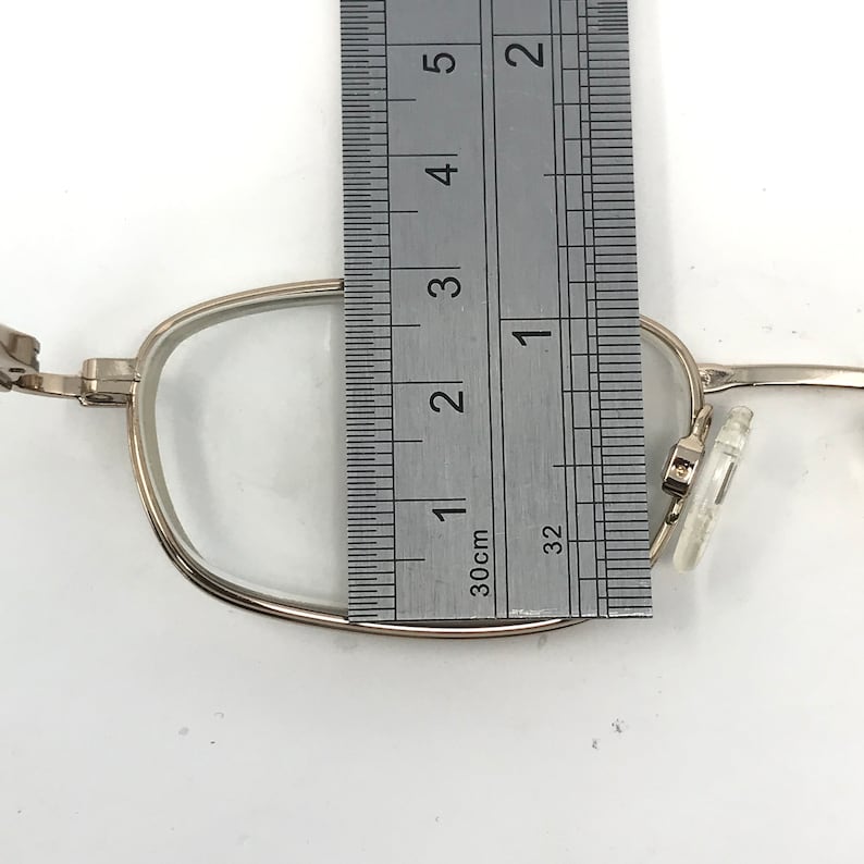 May include: Silver-framed eyeglasses with clear lenses are measured with a ruler. The frame is approximately 1 inch wide. The glasses are set against a white background, showing the details of the frame and lenses.