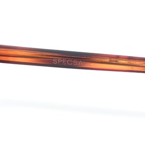 May include: Close-up of a brown eyeglasses frame with the text "SPECSAVERS" printed on the arm. The frame has a gradient color, transitioning from a lighter shade to a darker brown.