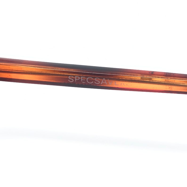 May include: Close-up of a brown eyeglasses frame with the text "SPECSAVERS" printed on the arm. The frame has a gradient color, transitioning from a lighter shade to a darker brown.