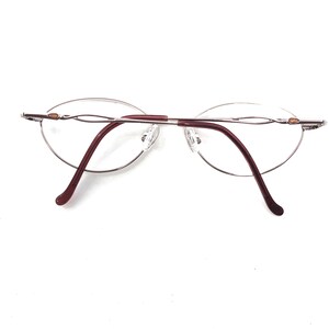 Puede incluir: Gafas sin montura con lentes ovaladas y montura de metal plateado. Las varillas son de color burdeos oscuro y las almohadillas nasales son transparentes. Las gafas est&aacute;n sobre un fondo blanco.