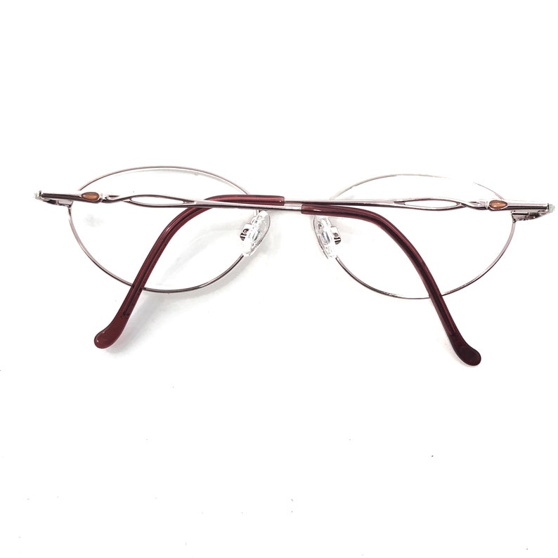 Puede incluir: Gafas sin montura con lentes ovaladas y montura de metal plateado. Las varillas son de color burdeos oscuro y las almohadillas nasales son transparentes. Las gafas est&aacute;n sobre un fondo blanco.