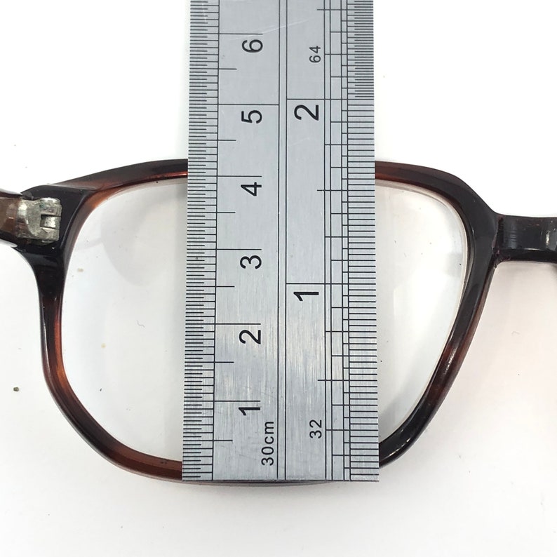 May include: Close-up of a pair of brown-rimmed eyeglasses with clear lenses. A metal ruler is placed over the glasses, measuring approximately 2 inches in height. The glasses have a classic, rectangular shape.